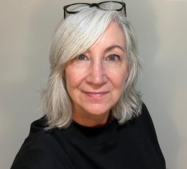 A medium close-up, front-facing portrait of a woman with shoulder-length, wavy silver hair and a slight smile. She wears a black top and black-rimmed glasses perched on top of her head. The background is a plain, neutral-toned wall. The lighting is soft and even.