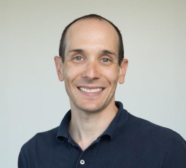 A head-and-shoulders portrait of a smiling man with a fair complexion, short brown hair, and green eyes. He is wearing a dark navy blue polo shirt and is positioned against a plain, light gray background. He has a friendly, approachable expression.