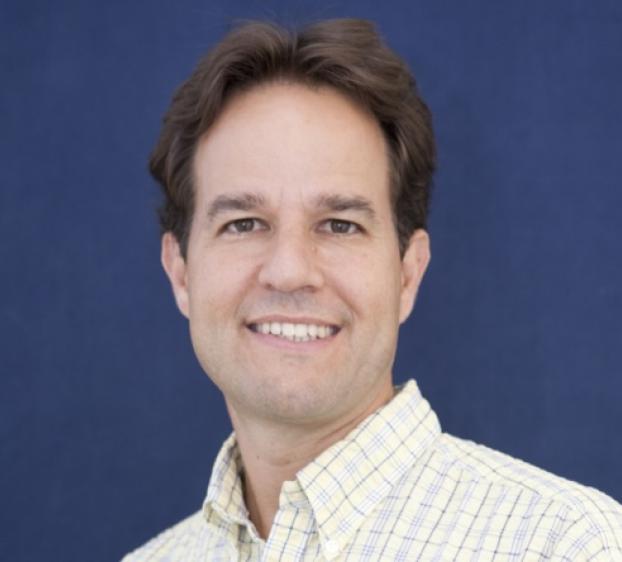 Headshot of a smiling man with brown hair and brown eyes, looking directly at the camera. He is wearing a light yellow and white checkered button-down shirt. The background is a solid, dark blue textured surface. The lighting is bright and even, highlighting his features.