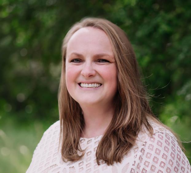 A headshot of a smiling woman with light brown hair and fair skin. She is wearing a cream-colored lace top with a diamond pattern. The background is a soft, blurred green of trees and foliage in natural daylight. She has a warm expression, looking directly at the camera.