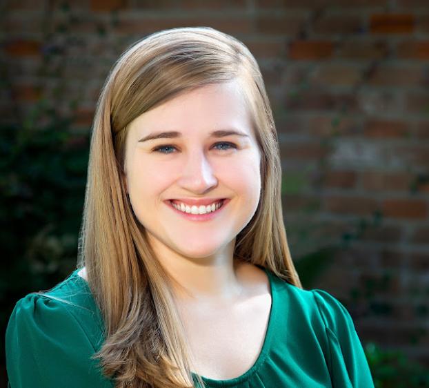 A professional headshot of a smiling woman with long blonde hair and blue eyes. She is wearing a dark green blouse and is positioned in front of a soft-focus background featuring a brick wall and green foliage. The lighting is bright and even, highlighting her friendly expression.