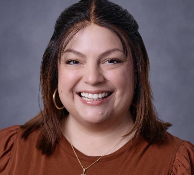 Headshot of a smiling woman with medium-length brown hair against a neutral gray background. She is wearing a rust-colored blouse, gold hoop earrings, and a gold necklace with a small cross pendant. She has a warm expression and her hair is parted in the middle with the front sections pulled back.