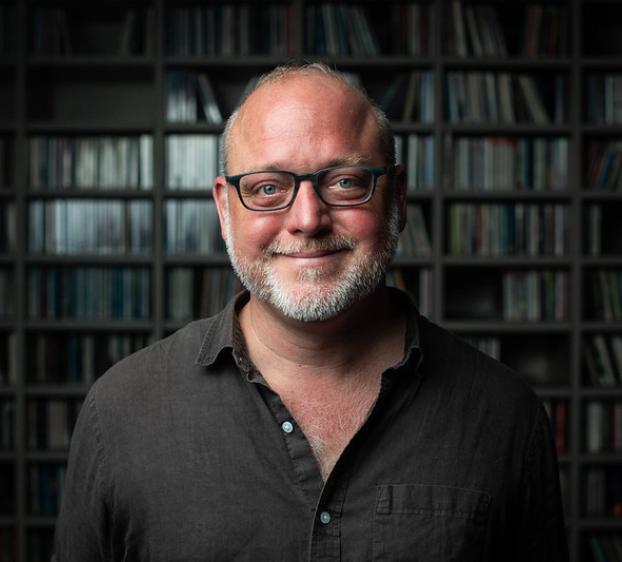 A headshot of a man with a fair complexion, short graying beard, and glasses. He is smiling warmly at the camera, wearing a dark button-down shirt. The background is a softly blurred, floor-to-ceiling bookshelf filled with numerous books, creating a scholarly and professional atmosphere.