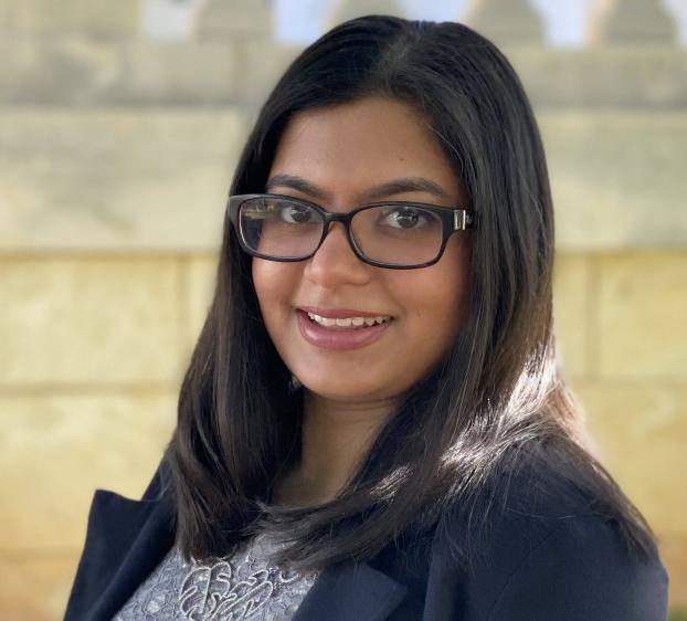 A headshot of a smiling young woman with long, dark hair and glasses. She is wearing a grey lace-patterned top underneath a dark blazer. The background is a soft-focus, light-colored stone wall, with natural sunlight highlighting the side of her hair.