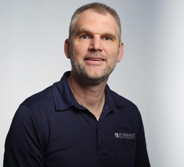 A professional headshot of a man with short, salt-and-pepper hair and a light beard. He is smiling slightly and wearing a dark blue polo shirt featuring the St. Edward’s University logo on the chest. The background is a clean, neutral light gray with soft studio lighting.