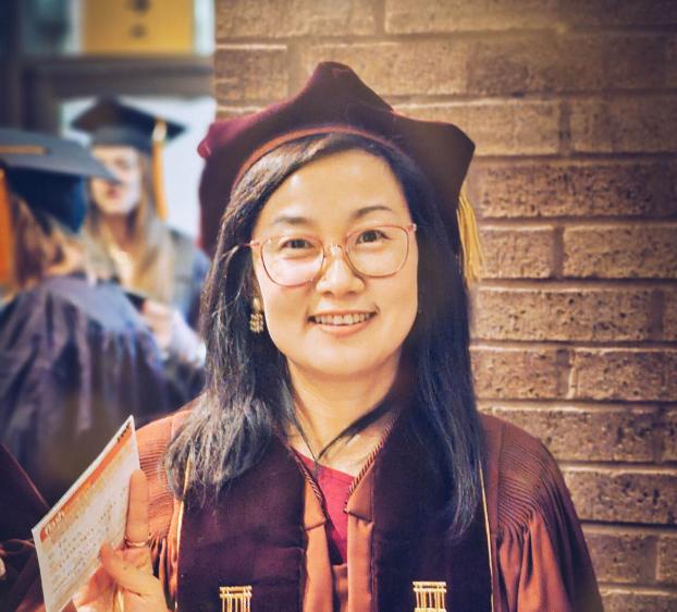 A smiling woman in her graduation regalia stands against a brick wall. She wears a maroon doctoral gown with gold velvet panels, a matching velvet tam, and pink-rimmed glasses. She holds a small card in one hand and a red and white stole over her other arm.