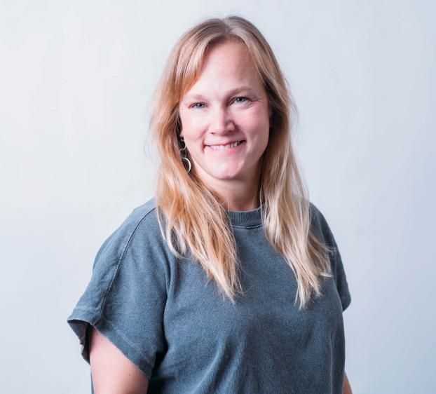 A medium shot of a smiling woman with long blonde hair and blue eyes, positioned against a plain, light grey background. She is wearing a charcoal grey, short-sleeved t-shirt and silver dangling earrings. She is angled slightly to the left while looking directly at the camera with a friendly expression. The lighting is bright and even, creating a professional yet casual headshot.