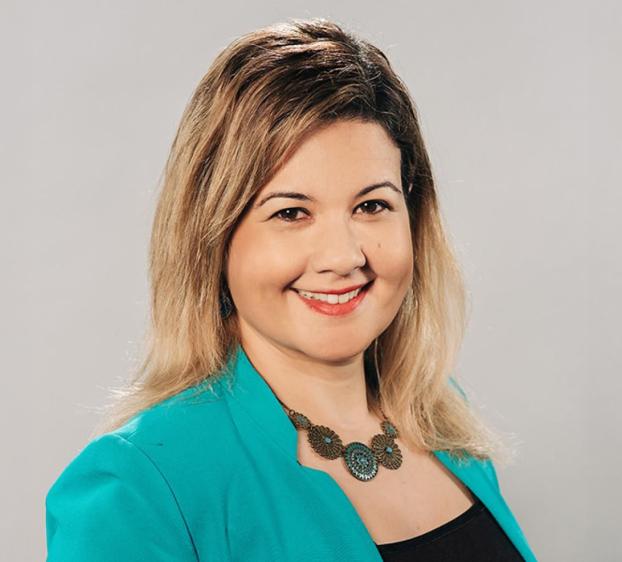 A professional headshot of a woman with blonde-highlighted hair, smiling warmly. She is wearing a vibrant turquoise blazer over a black top and a decorative turquoise and bronze statement necklace. The background is a clean, neutral light gray. The lighting is bright and even, highlighting her face and professional attire.