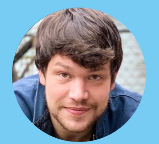 A circular headshot of a man with brown, wavy hair and a light beard, looking directly at the camera with a subtle smile. He is wearing a blue jacket. The portrait is set against a solid light-blue background.