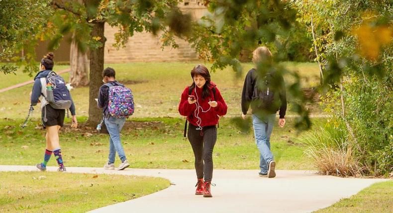 The image shows four students walking along a paved path in a park-like setting with green grass and trees. One student in the foreground is wearing a red jacket and red boots, looking at their phone with earphones on. Two students are walking away from the camera on the left, one wearing colorful socks and carrying a backpack, and the other wearing jeans and a colorful backpack. Another student on the right is also walking away, wearing a black hoodie and jeans.