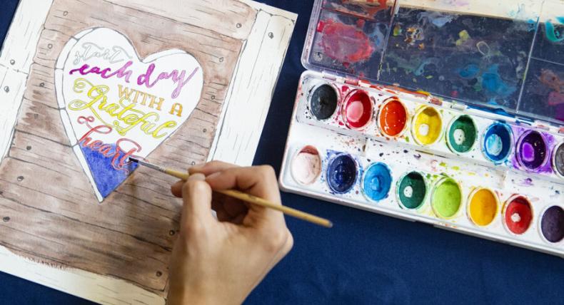 A person paints with watercolors on a page that reads "start each day with a grateful heart" and depicts a heart on wood.