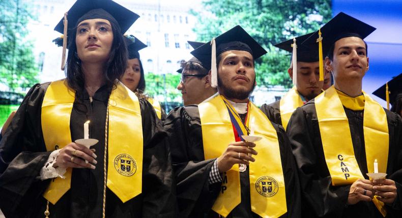 Three CAMP Students hold candles while wearing graduation caps, tassels, robes and stoles. 