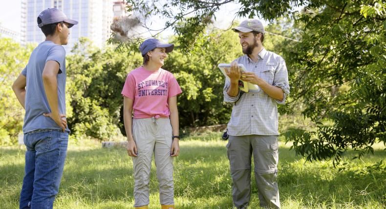 The image shows three people standing in a grassy outdoor area. The person on the left is wearing a blue t-shirt, jeans, and a cap, standing with hands on hips. The person in the middle, wearing a pink "St. Edward's University" t-shirt, beige pants, yellow boots, and a cap, is smiling at the person on the right. The person on the right, wearing a gray shirt, cargo pants, and a cap, is holding a tablet and smiling. The setting is sunny with green trees and a building visible in the background.