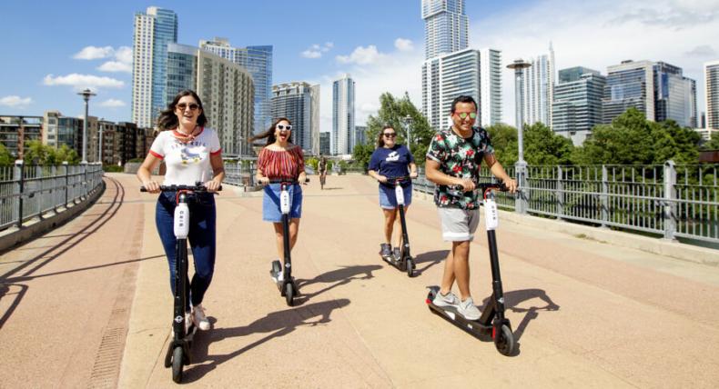 Four students ride scooters acros the Pfluger Pedestrian Bridge with the skyline of Austin in the background.