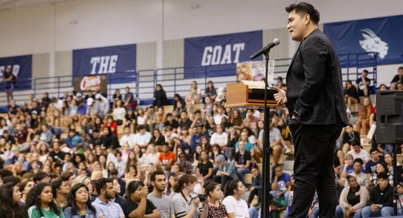 Jose Antonio Vargas stands in front of a podium and microphone and speaks to the audience in the RAC gym.