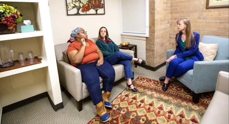 The image depicts three individuals seated in a room. On the left, a person wearing blue jeans and an orange top sits on a beige sofa In the middle, another individual in green pants and a red top occupies a chair with green upholstery. The person on the right, wearing dark pants and a blue top, sits on a light blue armchair. The room features decorative elements like paintings, flowers, and a patterned area rug.