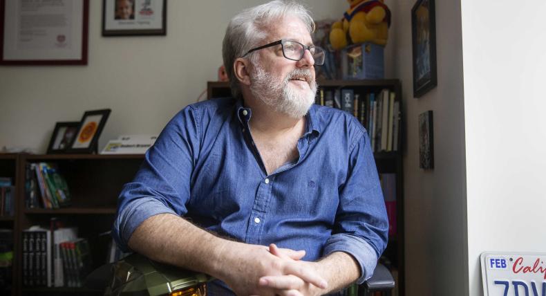 The image shows a man with gray hair and a beard, wearing glasses and a blue button-up shirt, sitting in a room. He is looking to the side with a thoughtful expression. Behind him, there are bookshelves filled with books and various items, including a stuffed Winnie the Pooh bear wearing a cape. On the wall, there are framed pictures and a sign that reads "Events" with a name and photo. The setting appears to be an office or study.