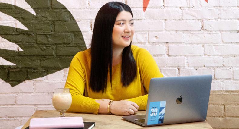 The image depicts an individual sitting at a wooden table with a laptop open in front of them. The laptop displays a blue graph or chart. To the left is a pink notebook, and in front of the person is a glass containing a creamy beverage. The person wears a yellow long-sleeved top. Behind them is an interior wall with a green artistic design or logo.