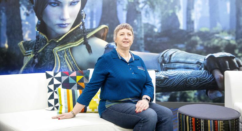 The image shows a woman sitting on a white couch in front of a large poster or screen displaying a character from a video game. The woman sitting on the couch is dressed in a blue checkered shirt and jeans, with short hair. She is smiling slightly and appears to be in a relaxed pose. There are colorful pillows on the couch, and a small round stool or table with a patterned cover is positioned next to the couch. The setting appears to be casual and modern, likely within a gaming or tech environment.