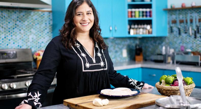 The image features a woman with long, dark hair, wearing a black blouse with white embroidery. She is standing in a bright kitchen with turquoise cabinets and a mosaic tile backsplash. She is smiling and appears to be preparing food, as there is a cutting board with dough and a plate with tortillas in front of her. A basket of vegetables and a tortilla press are also visible on the counter.