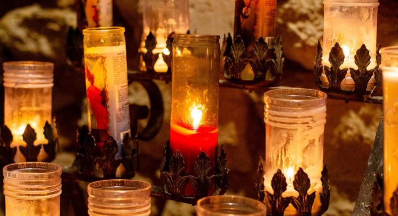 The image shows a collection of candles placed within glass holders. The candles vary in height and the amount of wax melted, suggesting they have been burning for different durations. Some candles are lit, casting a warm, inviting glow, while others remain unlit. The glass holders feature decorative bases, possibly made of metal with intricate designs. The background is blurred but appears to be a wall with patterns, likely reflections or shadows created by the candlelight.