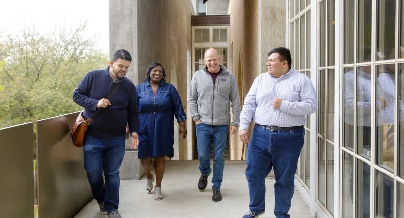 The image shows four people walking together along an outdoor corridor with large windows on the right and a railing on the left. The group consists of two men and two women, all casually dressed and engaged in conversation, creating a friendly and collegial atmosphere. The background includes greenery visible through the windows and an architectural setting with concrete and brick elements. The scene conveys a sense of collaboration and camaraderie.