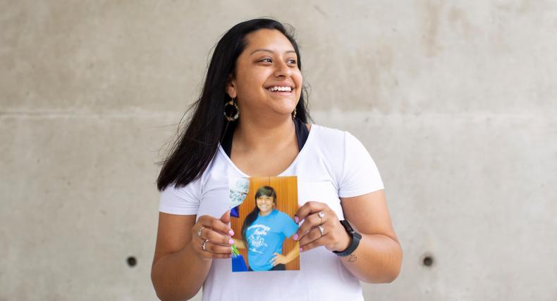 The image shows a young woman standing against a concrete wall. The woman is wearing a white t-shirt with some visible graphic designs on it. The person is holding a photograph that shows another individual wearing a blue t-shirt with some text or design on it. 