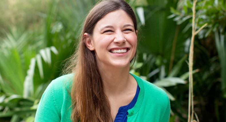 The image shows an individual standing in front of a lush background filled with green foliage. The person has long, light brown hair and is wearing a bright green top paired with a blue cardigan.