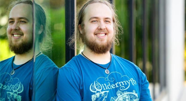 The image shows a bearded man with long hair, smiling while standing next to a reflective glass surface. He is wearing a blue T-shirt with the word "Wildermyth" and a detailed design on it. The reflection in the glass creates a mirrored effect, showing both his front and side profile. The background is blurred, suggesting an outdoor setting with natural light. He is also wearing a necklace with a circular pendant.