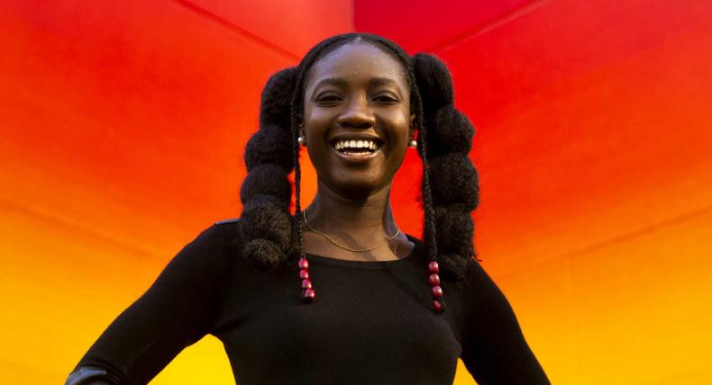 Kadija Samura smiles and stands in front of a warm gradient mural.