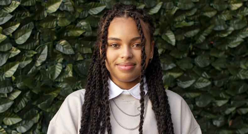 Taylor Huey wears a neutral tan sweatshirt with a white color shirt and multiple necklaces and stands in front of a green ivy background.