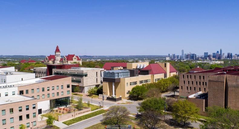 he image depicts an aerial view of a campus with various buildings. In the foreground, multi-story buildings with distinct architectural designs are visible, including one with a prominent red roof. The campus is well-maintained, surrounded by green spaces. The clear sky above and distant city skyline suggest an urban location. No people or vehicles are visible, creating a serene atmosphere. This image likely showcases the layout and proximity of an educational institution to the nearby city.