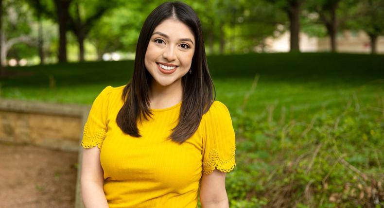 Victoria Garcia wears a yellow shirt and sits on a bench on Ragsdale patio. Greenery and trees are in the background.