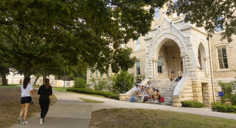 Two students walk on a path as other students sit on the steps of Holy Cross Hall.