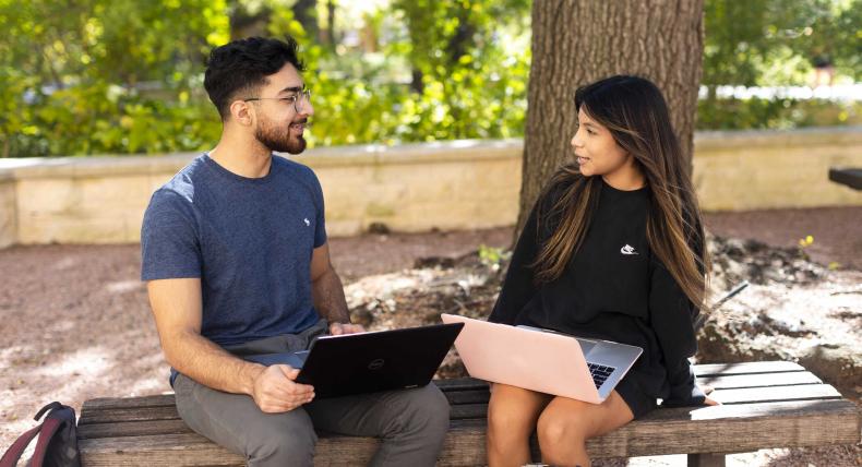  The image depicts two individuals sitting on a bench outdoors. Both are using laptops—one person wears a blue t-shirt, while the other wears a black long-sleeve shirt. They appear focused on their screens, suggesting work or study. The setting includes trees, creating a peaceful atmosphere.