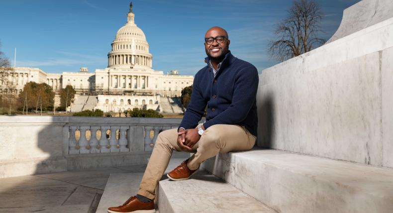 The image features a man sitting on marble steps with the United States Capitol building prominently in the background. He is smiling, wearing a dark blue cardigan, beige pants, and brown shoes. The setting is outdoors under a clear blue sky. The composition highlights both the individual and the iconic architecture, suggesting a professional or formal context. The overall atmosphere is bright and positive.