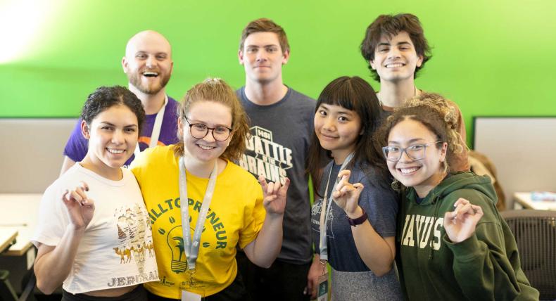 Seven students stand in front of a green wall and give toppers up hand signs.