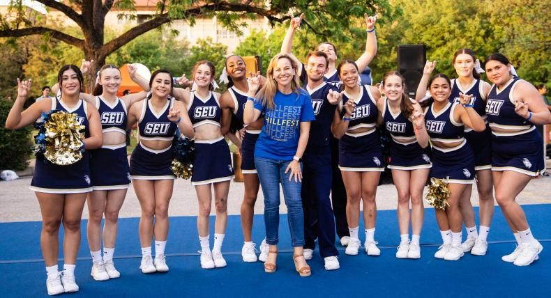 The image shows a group of cheerleaders and a few other individuals posing together outdoors. They are wearing matching navy blue and white uniforms with "SEU" printed on them. One person in the middle is wearing a blue T-shirt with "HILLFEST" written on it. Everyone is smiling and making a hand gesture, indicating a spirited and joyful atmosphere. The background features trees and a building, suggesting a campus or park setting.