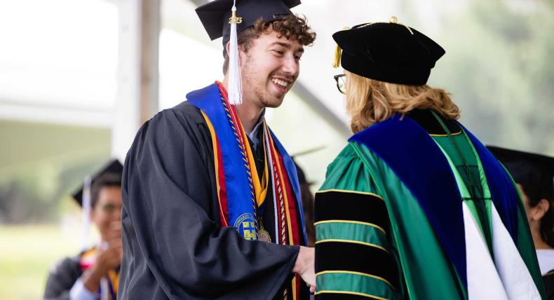 August Railey shakes hands with dean Catherine Campbell at commencement. Both are wearing caps and gowns for their commencement regalia.