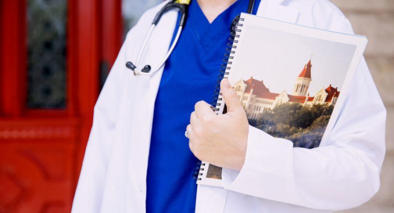 A person stands in front of hte red doors wearing blue scrubs a white coat and a stethoscope