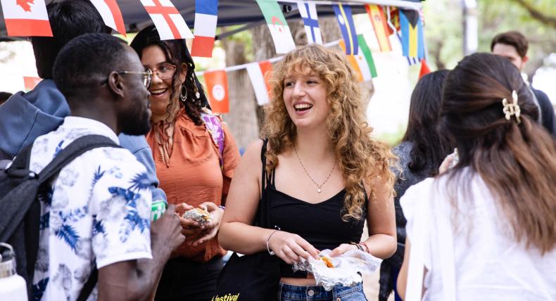 Melody Borg, center, smiles and talks with other international students at an event. Flags of different countries hang on a tent in the background.