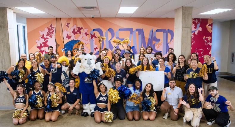 The image depicts a large group of people, including cheerleaders, faculty, and a mascot, posing enthusiastically in front of a colorful mural with the word "DREAMER" prominently displayed. The group is dressed in blue and gold, with many holding pom-poms, and includes the SEU (St. Edward's University) mascot. The atmosphere is festive and celebratory, suggesting a school or community event.