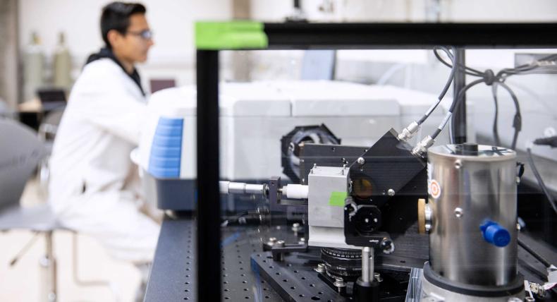 A student in a lab coat is in the background as a piece of research equipment is in focus in the foreground.