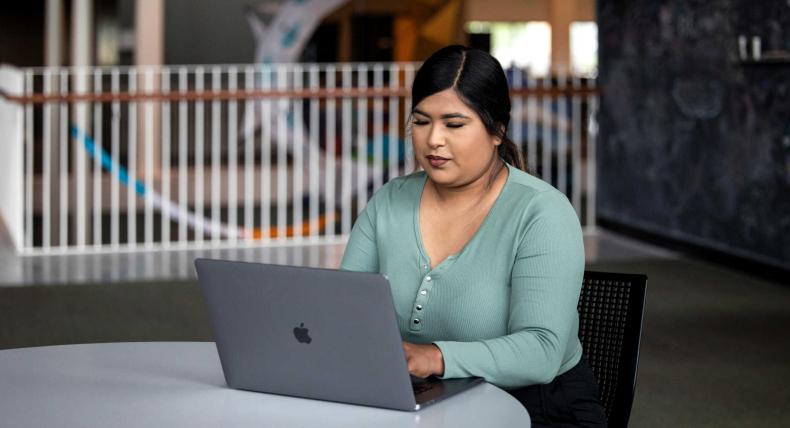 A student sits at a desk in the library typing on their laptop.