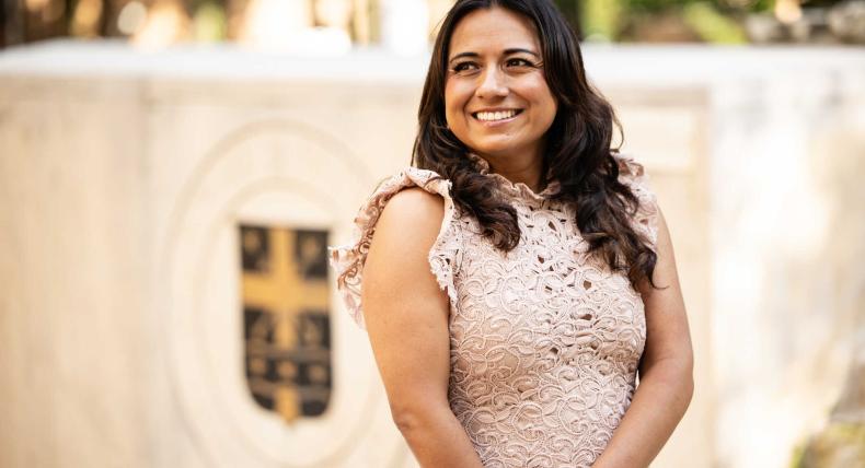 The image shows a woman with long dark hair wearing a light pink lace dress, smiling warmly while standing outdoors. In the background, there is a blurred emblem or crest on a stone structure. The setting appears to be a formal or professional environment, possibly a campus or an institution. The woman looks composed and confident.