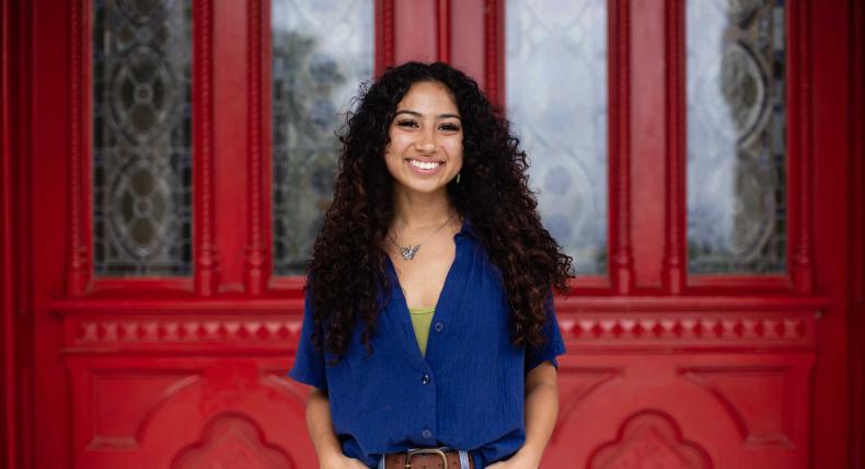 A young woman with curly hair stands smiling in front of an ornate red door with decorative glass panels. She is wearing a blue button-up shirt over a green top, paired with denim jeans. Her hands are in her pockets, and she is accessorized with a butterfly necklace. The vibrant red of the door contrasts with her outfit, creating a striking background for the portrait.