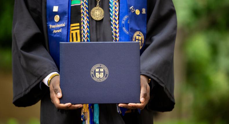 A student adorned with stoles and cords in their cap and gown holds their degree.