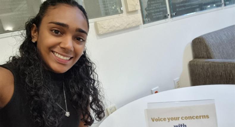 Analee Maharaj wears a black blouse and necklace and takes a selfie while sitting at a white table.