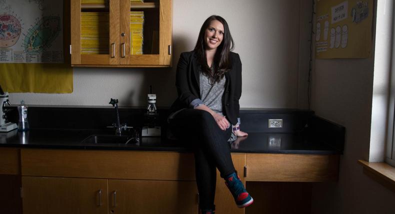 The image shows a woman sitting on a black countertop in a laboratory or classroom setting. She is smiling and casually dressed in a gray shirt, black blazer, and black pants with colorful sneakers. Behind her are wooden cabinets, a poster depicting animal and plant cells, and a microscope on the counter. The lighting is soft, with natural light coming from a window on the right, creating a relaxed and approachable atmosphere.