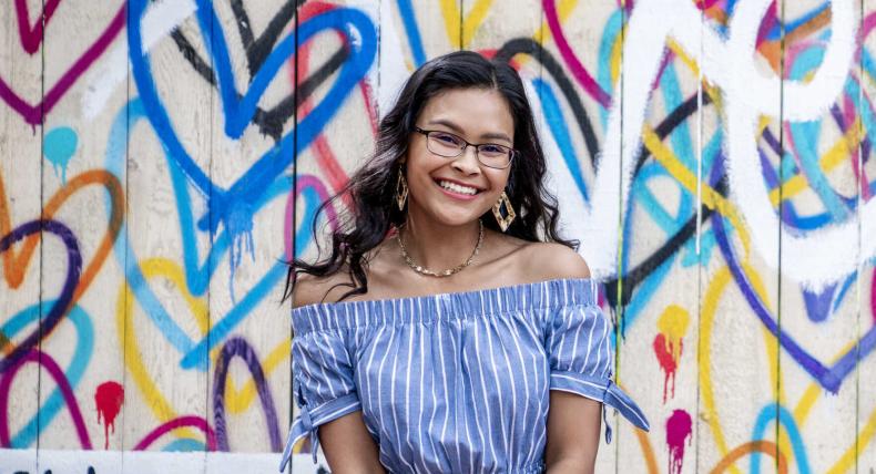 The image features a young woman with long, dark, wavy hair wearing glasses. She is smiling and dressed in an off-the-shoulder, blue and white striped top with short sleeves tied at the ends. She accessorizes with large, geometric earrings and a delicate necklace. The background is a vibrant mural with colorful heart shapes and abstract designs, adding an artistic and lively atmosphere to the image.